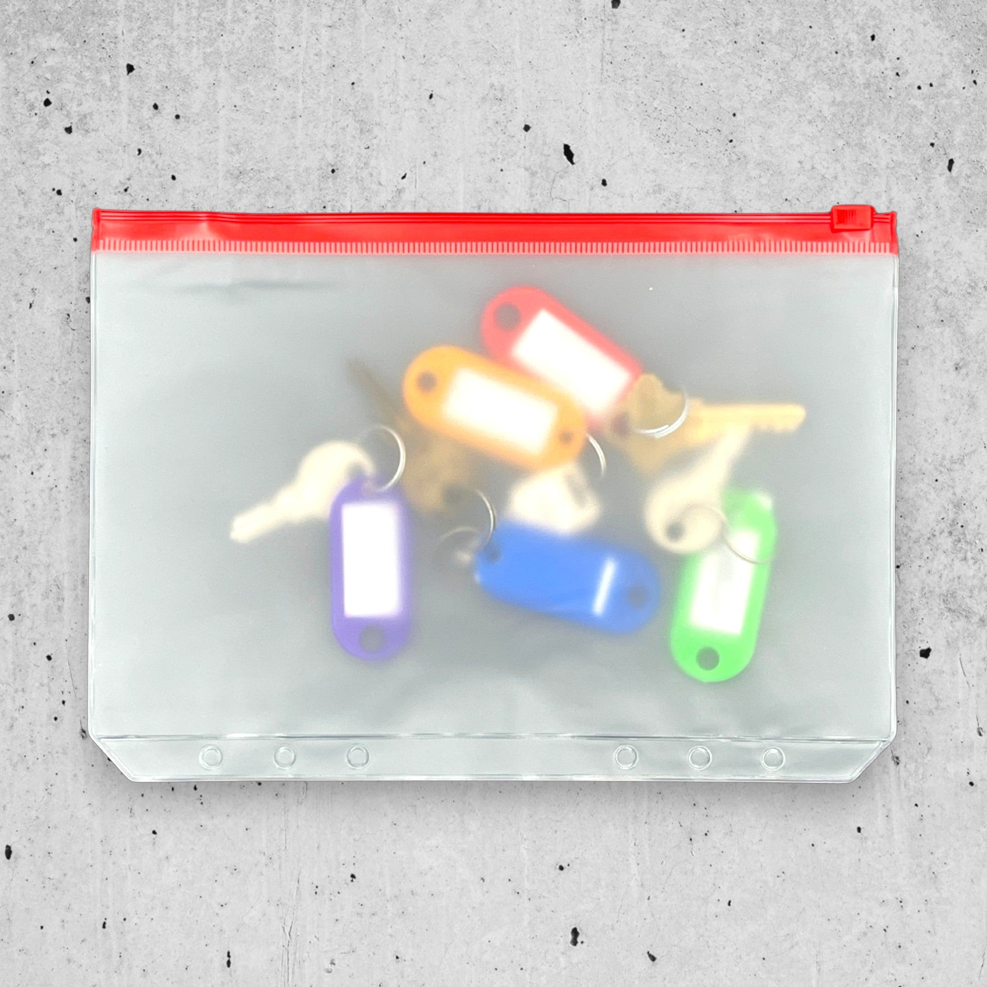Colorful key tags inside a clear pouch with red zipper on a gray concrete surface